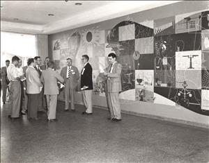 Several men in suits stand in front of a complex mural containing planets, human bodies, graphs, equations, and other symbols of science. 